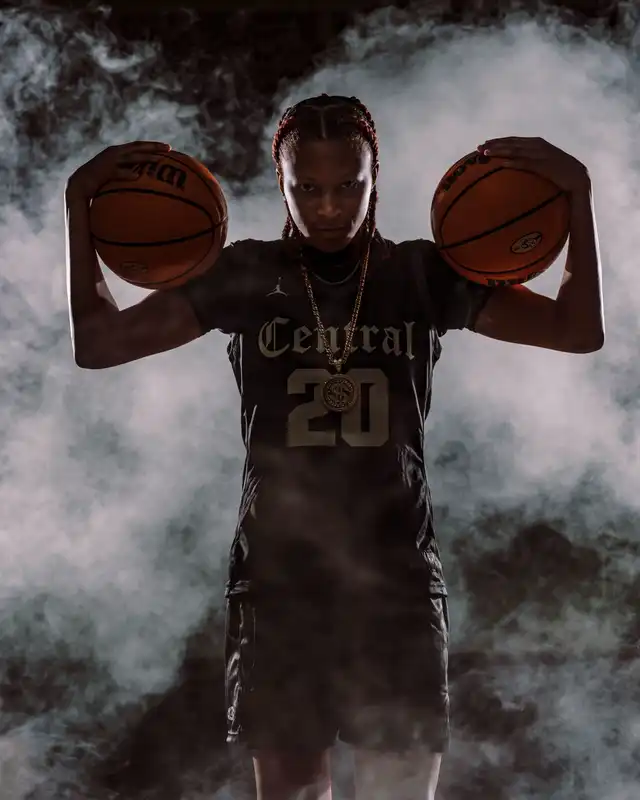 A female basketball player in a dark jersey with 'Central' and number 20 holds two sports balls near her head. She wears a chain, standing amidst smoke with an intense expression.